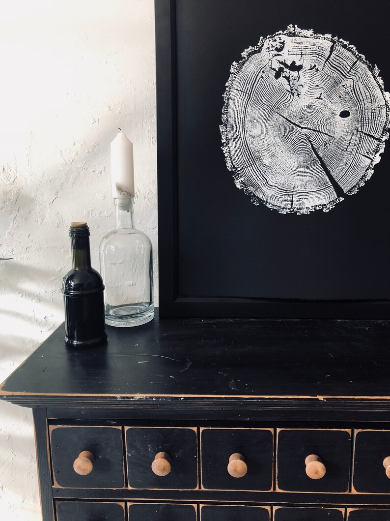 May include: A black dresser with multiple drawers, each with a wooden knob. On top of the dresser is a framed black and white print of a tree trunk cross-section. There are also two glass bottles and a wine bottle on the dresser.