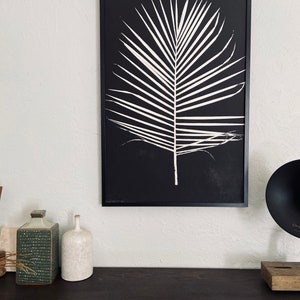 May include: A black and white framed print of a palm leaf on a black background. The print is hanging on a white wall above a dark brown wooden surface.