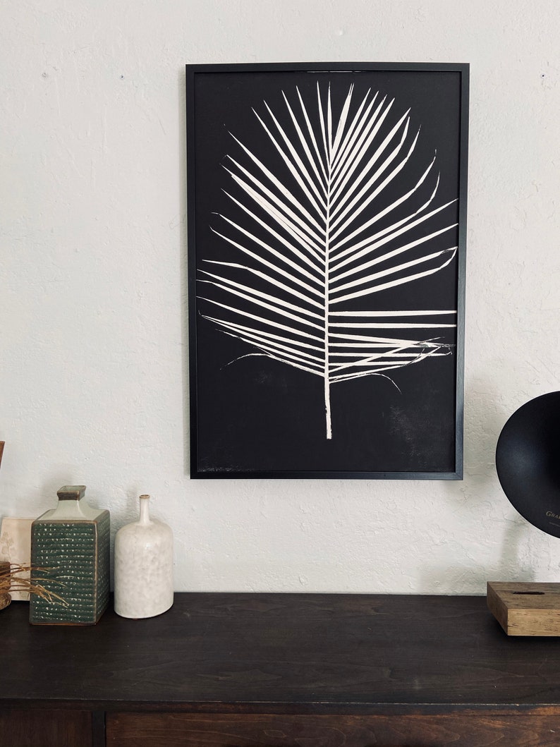 May include: A black and white framed print of a palm leaf on a black background. The print is hanging on a white wall above a dark brown wooden surface.