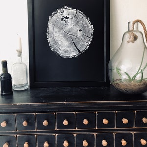 May include: A black framed print of a white tree ring on a black background. The print is sitting on a black dresser with multiple drawers. The dresser has wooden knobs.