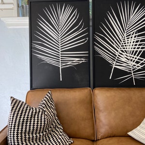May include: Two black framed prints of white palm leaves on a brown leather couch with a black and white patterned throw pillow.