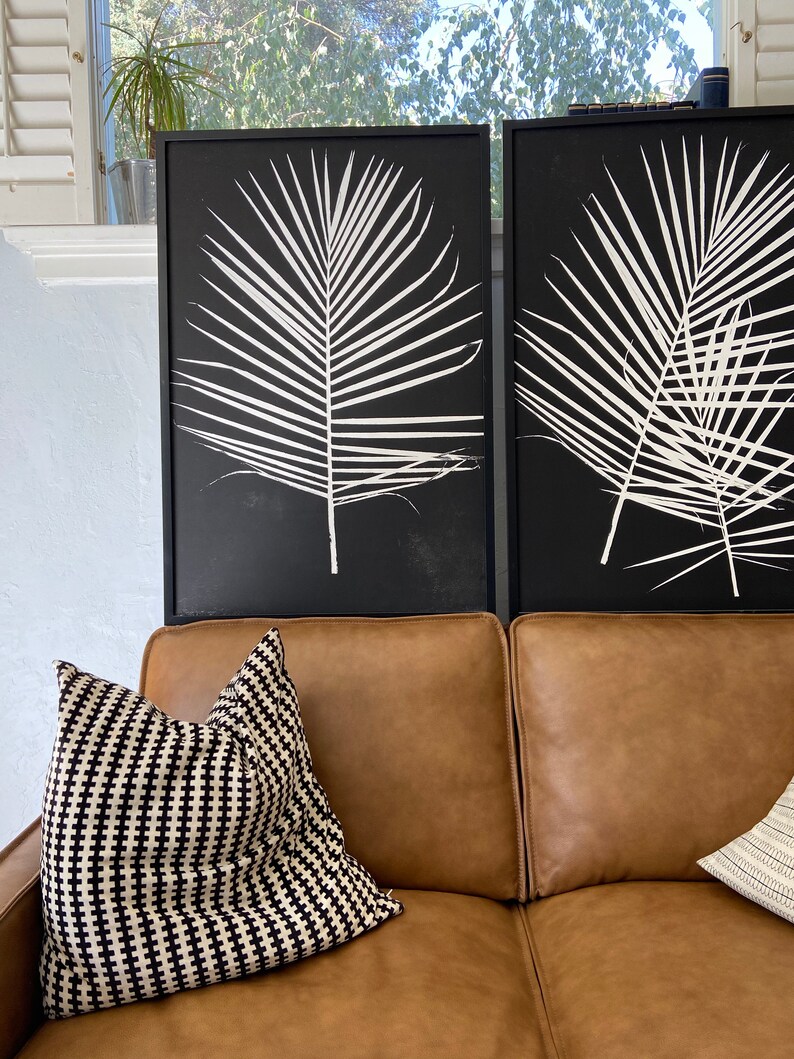 May include: Two black framed prints of white palm leaves on a brown leather couch with a black and white patterned throw pillow.