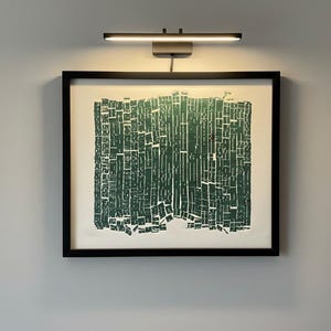 Puede incluir: Una impresión artística abstracta enmarcada con un diseño geométrico verde y blanco. La obra de arte está montada en un marco negro e iluminada por una pequeña lámpara de cuadro rectangular plateada.