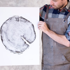 Op de afbeelding: Een zwart-witte print van een dwarsdoorsnede van een boomstam. De print is op een witte achtergrond en wordt vastgehouden door een persoon die een bruine schort en een geruit overhemd draagt.