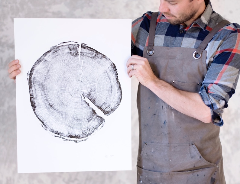 Op de afbeelding: Een zwart-witte print van een dwarsdoorsnede van een boomstam. De print is op een witte achtergrond en wordt vastgehouden door een persoon die een bruine schort en een geruit overhemd draagt.
