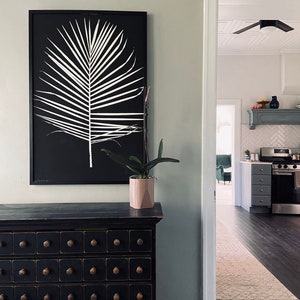 May include: A black framed print of a white palm leaf on a black background hangs above a black dresser with multiple drawers. A potted plant sits on the dresser.