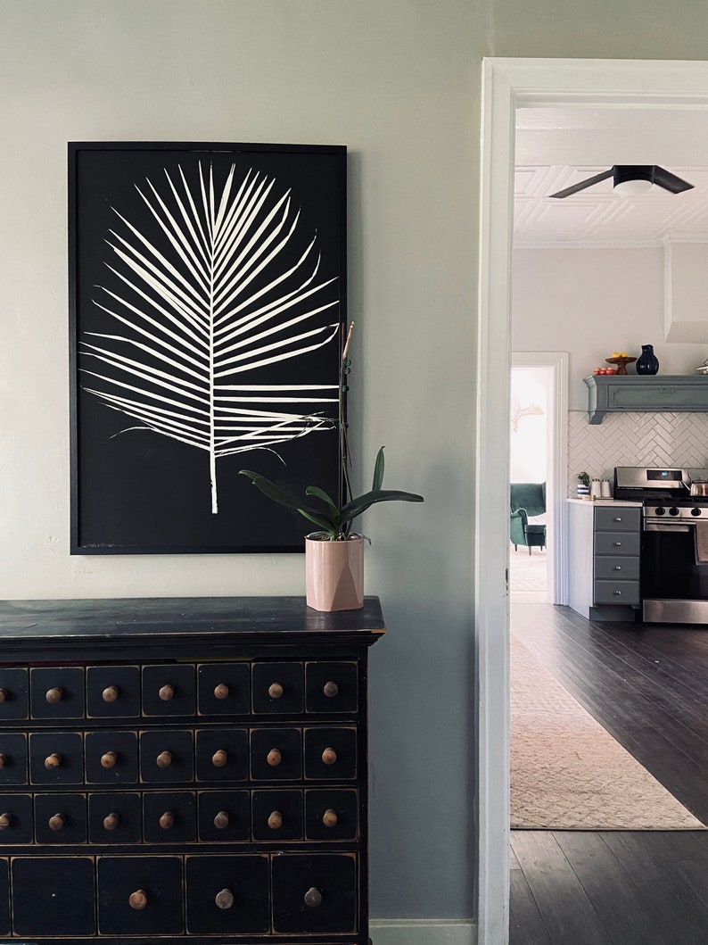 May include: A black framed print of a white palm leaf on a black background hangs above a black dresser with multiple drawers. A potted plant sits on the dresser.