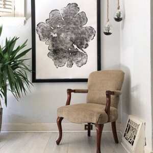 May include: A black and white print of a tree trunk cross-section, framed in black, hangs on a white wall above a beige armchair with wooden legs.