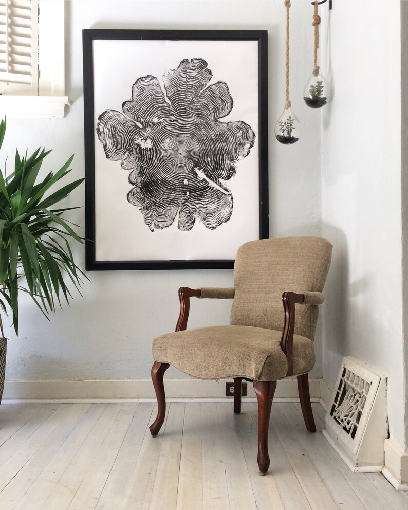 May include: A black and white print of a tree trunk cross-section, framed in black, hangs on a white wall above a beige armchair with wooden legs.