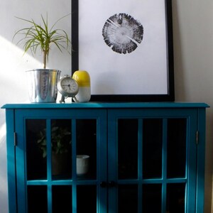 May include: A teal blue cabinet with glass doors and shelves. The cabinet has a black clock and a yellow and white ceramic vase on top. A framed black and white print of a tree trunk cross section is hanging on the wall above the cabinet.