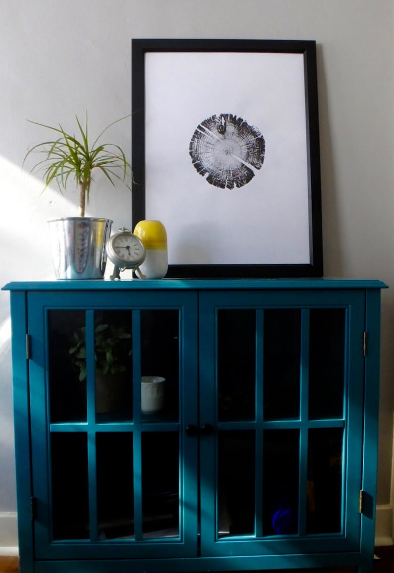 May include: A teal blue cabinet with glass doors and shelves. The cabinet has a black clock and a yellow and white ceramic vase on top. A framed black and white print of a tree trunk cross section is hanging on the wall above the cabinet.