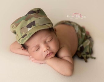 Puede incluir: Un bebé recién nacido durmiendo con una gorra de camuflaje de estilo militar y una braga a juego. El bebé está acostado boca abajo con los brazos cruzados, descansando sobre una superficie de color crema. La gorra y la braga presentan un patrón de camuflaje verde, marrón y beige.