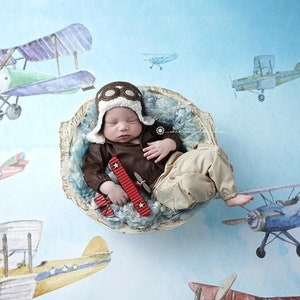May include: A newborn baby wearing a brown leather aviator costume and a brown leather aviator hat with goggles, sleeping in a white bowl with blue and white fabric. The baby is holding a red toy airplane. The background is a light blue watercolor with images of airplanes.