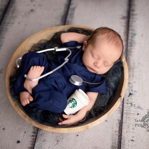 May include: A newborn baby wearing a blue scrubs outfit and a stethoscope, sleeping in a wooden bowl with a toy coffee cup.