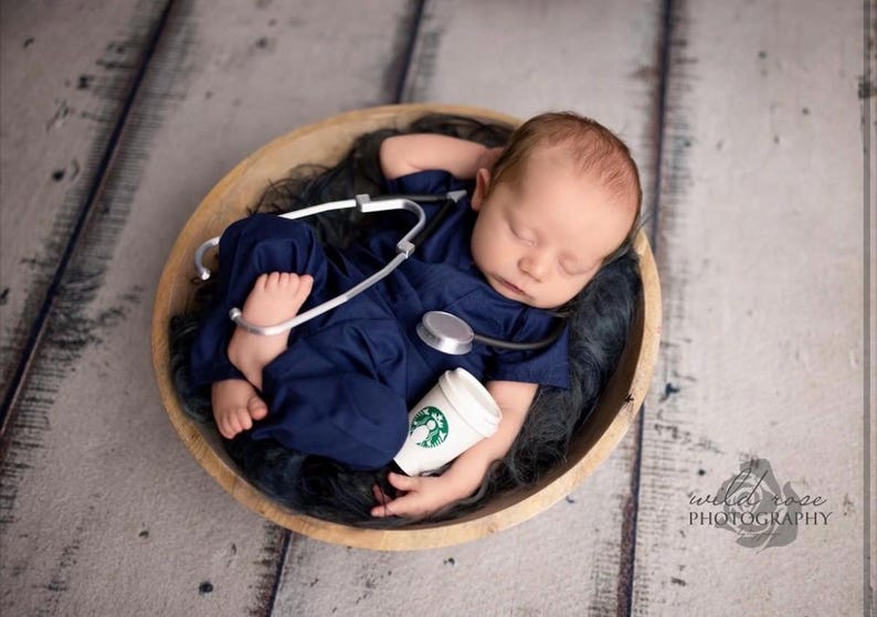 May include: A newborn baby wearing a blue scrubs outfit and a stethoscope, sleeping in a wooden bowl with a toy coffee cup.