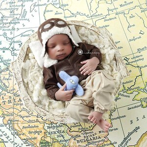May include: A baby wearing a brown leather aviator costume with a white fur-trimmed hat and goggles, holding a blue toy airplane. The baby is lying in a white, fluffy nest on top of a vintage world map.