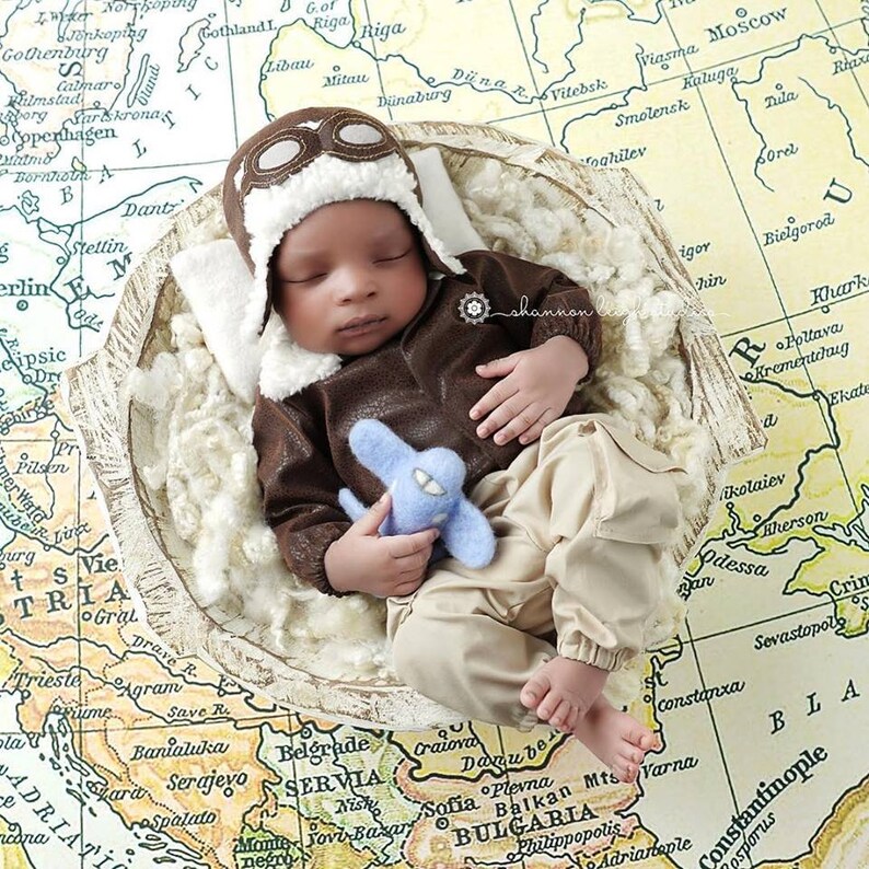 May include: A baby wearing a brown leather aviator costume with a white fur-trimmed hat and goggles, holding a blue toy airplane. The baby is lying in a white, fluffy nest on top of a vintage world map.