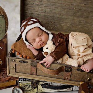 May include: A baby wearing a brown aviator hat and brown and beige clothing is sleeping in a wooden suitcase with a teddy bear. The suitcase is on top of other suitcases and a globe.