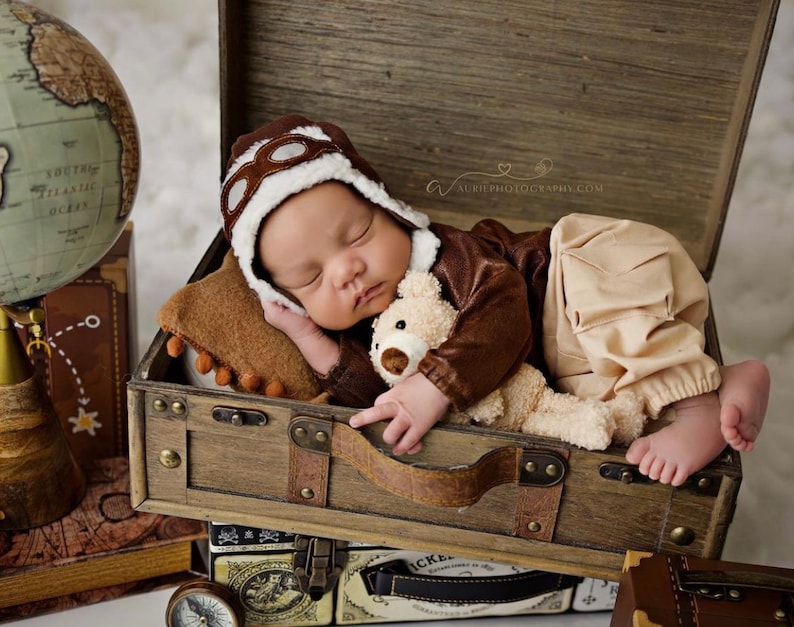 May include: A baby wearing a brown aviator hat and brown and beige clothing is sleeping in a wooden suitcase with a teddy bear. The suitcase is on top of other suitcases and a globe.