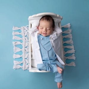 May include: A baby dressed in a white doctor's coat and blue scrubs with a stethoscope around their neck is lying in a white crib with a white crocheted rug underneath.