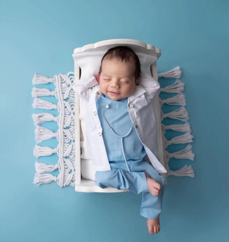 May include: A baby dressed in a white doctor's coat and blue scrubs with a stethoscope around their neck is lying in a white crib with a white crocheted rug underneath.