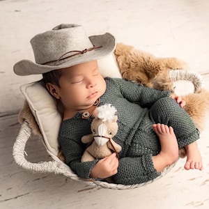 May include: A sleeping baby wearing a gray cowboy hat and a dark green knitted outfit, nestled in a woven basket with a plush toy horse. The baby is lying on a soft, furry blanket.