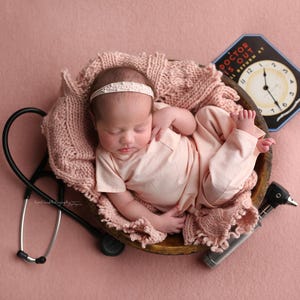 May include: A newborn baby wearing a pink outfit and a white headband is sleeping in a wooden bowl lined with a pink knitted blanket. A stethoscope, an otoscope, and a vintage clock with the text "Doctor Is Out" are on a pink background.