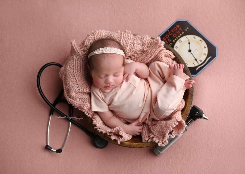 May include: A newborn baby wearing a pink outfit and a white headband is sleeping in a wooden bowl lined with a pink knitted blanket. A stethoscope, an otoscope, and a vintage clock with the text "Doctor Is Out" are on a pink background.