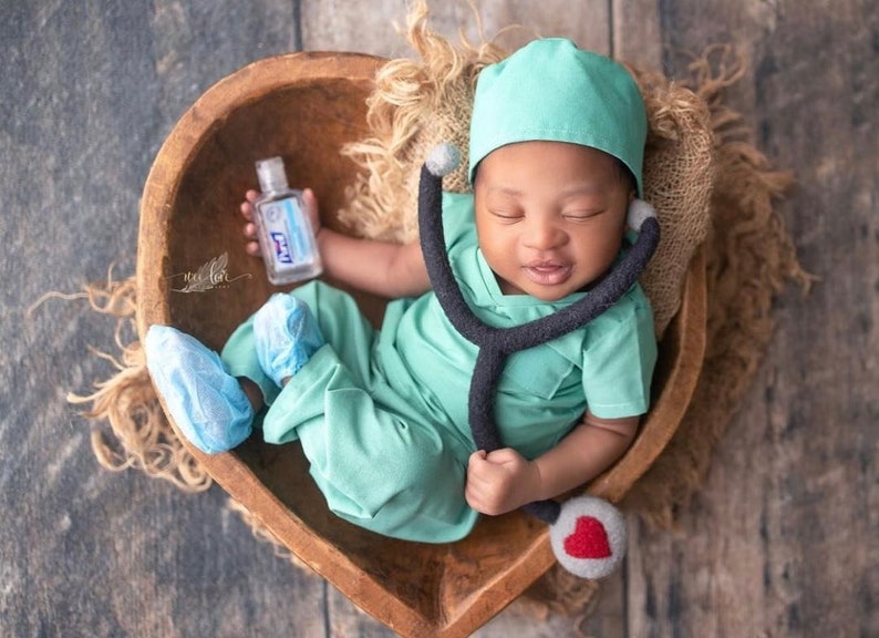 May include: A newborn baby dressed in a green medical scrubs outfit with a stethoscope and a small bottle of hand sanitizer. The baby is lying in a wooden bowl with a brown burlap background.