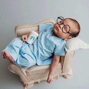May include: A baby wearing a light blue scrubs outfit and oversized black glasses is lying on a cream-colored couch. The baby is holding a small white cup with a green Starbucks logo.
