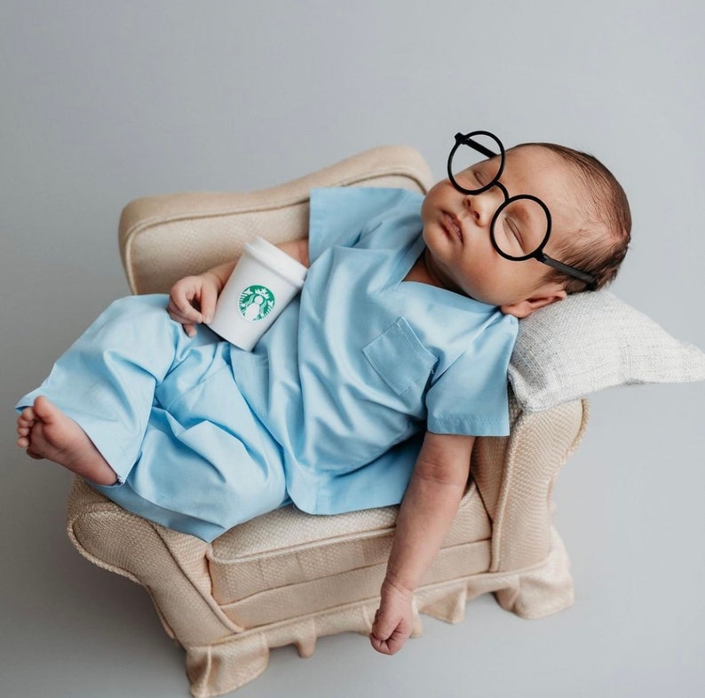 May include: A baby wearing a light blue scrubs outfit and oversized black glasses is lying on a cream-colored couch. The baby is holding a small white cup with a green Starbucks logo.