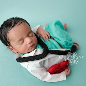 May include: A newborn baby wearing a white doctor's coat with a black stethoscope and a red heart. The baby is lying on a turquoise blue background.
