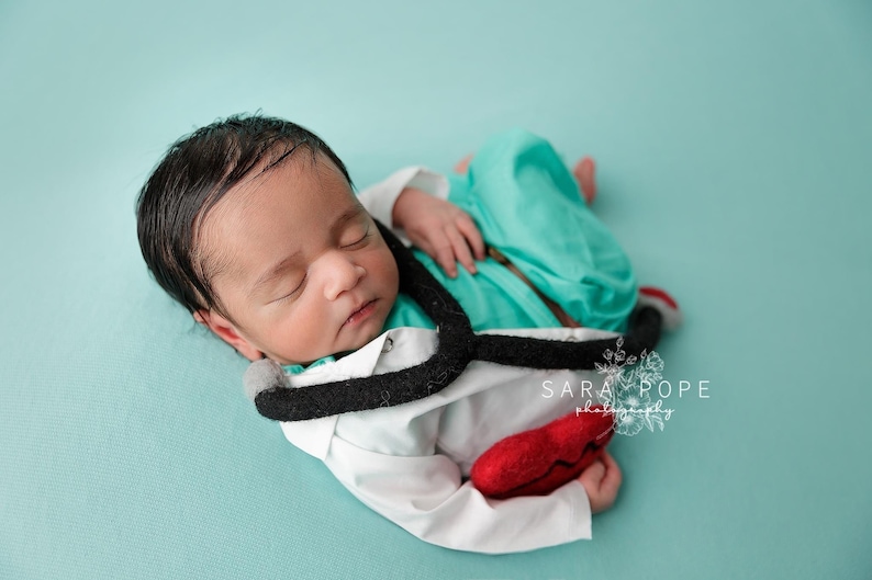 May include: A newborn baby wearing a white doctor's coat with a black stethoscope and a red heart. The baby is lying on a turquoise blue background.