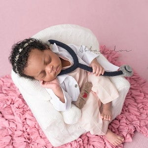 May include: A newborn baby wearing a white doctor's coat and a stethoscope, sleeping in a white knitted chair. The baby is wearing a headband with white beads and a pink and brown outfit. The chair is sitting on a pink ruffled blanket.