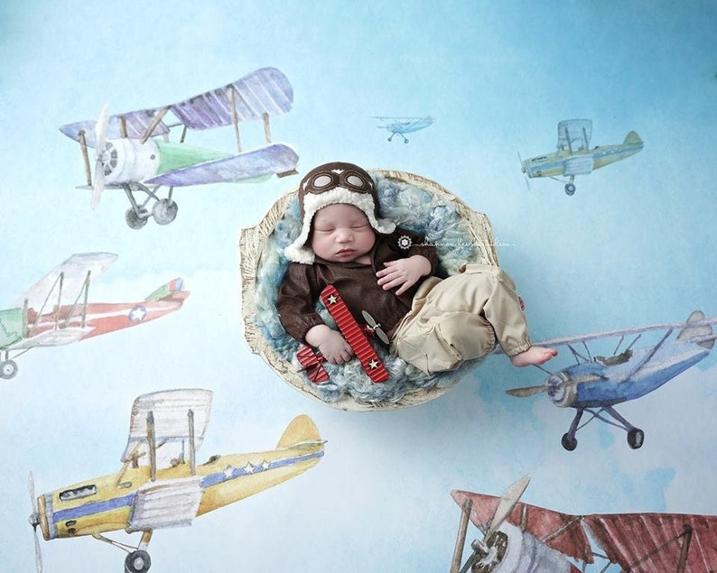May include: A newborn baby wearing a brown leather aviator costume and a brown leather aviator hat with goggles, sleeping in a white bowl with blue and white fabric. The baby is holding a red toy airplane. The background is a light blue watercolor with images of airplanes.