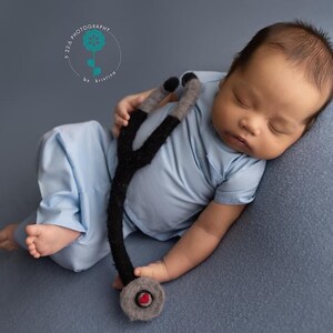 May include: A baby wearing a blue scrubs outfit and holding a black and gray toy stethoscope. The baby is sleeping on a gray background.