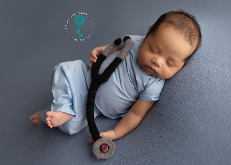 May include: A baby wearing a blue scrubs outfit and holding a black and gray toy stethoscope. The baby is sleeping on a gray background.