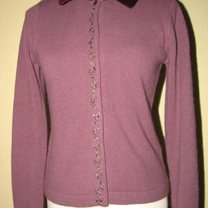 Puede incluir: Un c&aacute;rdigan malva con cuello de terciopelo y adornos plateados en la cremallera delantera. El cuello tiene un dise&ntilde;o de cuentas.