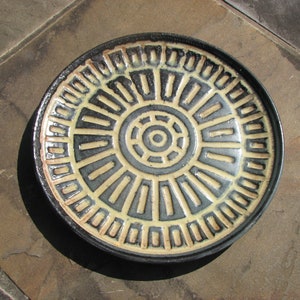 May include: A black and cream ceramic plate with a circular design. The design features concentric circles and radiating lines.