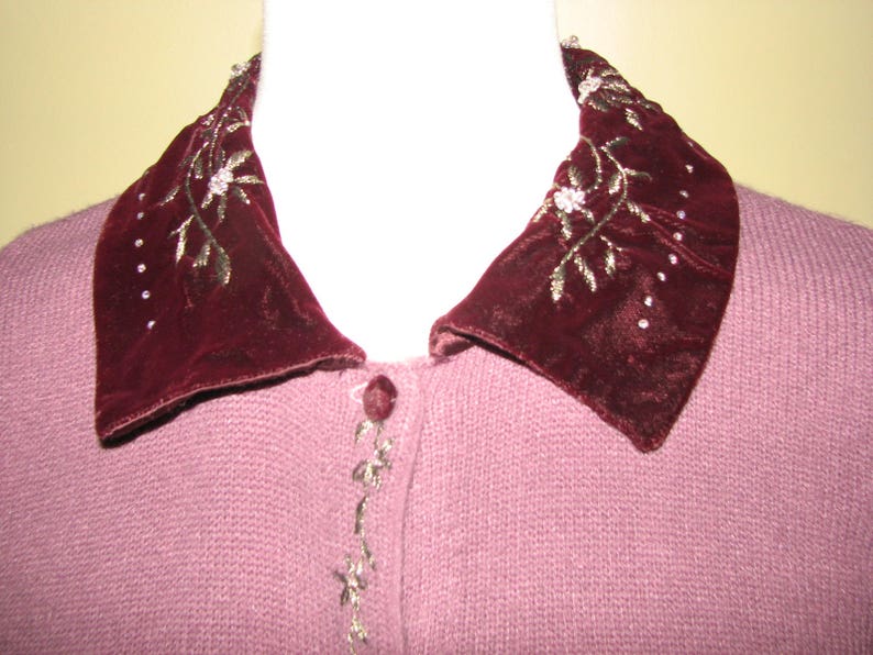 Puede incluir: Un c&aacute;rdigan de punto malva con un cuello grande de terciopelo burdeos con detalles florales bordados en hilo dorado.