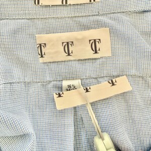 May include: Close-up of a light blue button-down shirt with a cream-colored label featuring the letter "T" repeated. Scissors are cutting the label. The shirt size is 16 1/2 34/35.