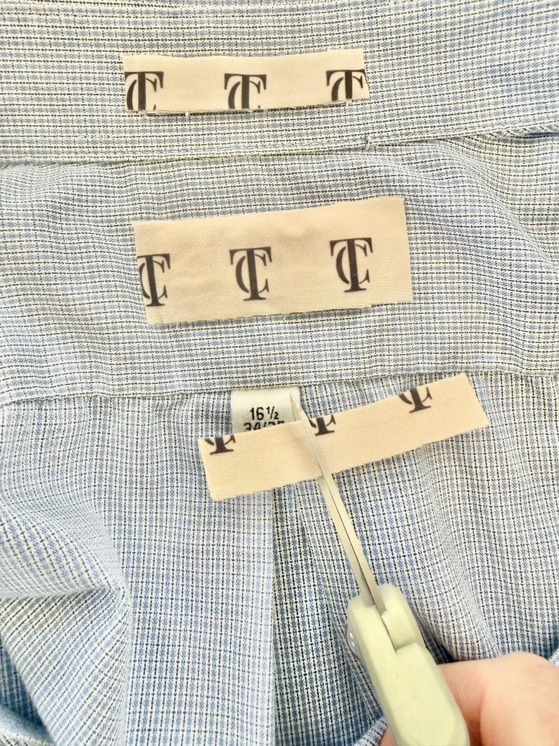 May include: Close-up of a light blue button-down shirt with a cream-colored label featuring the letter "T" repeated. Scissors are cutting the label. The shirt size is 16 1/2 34/35.