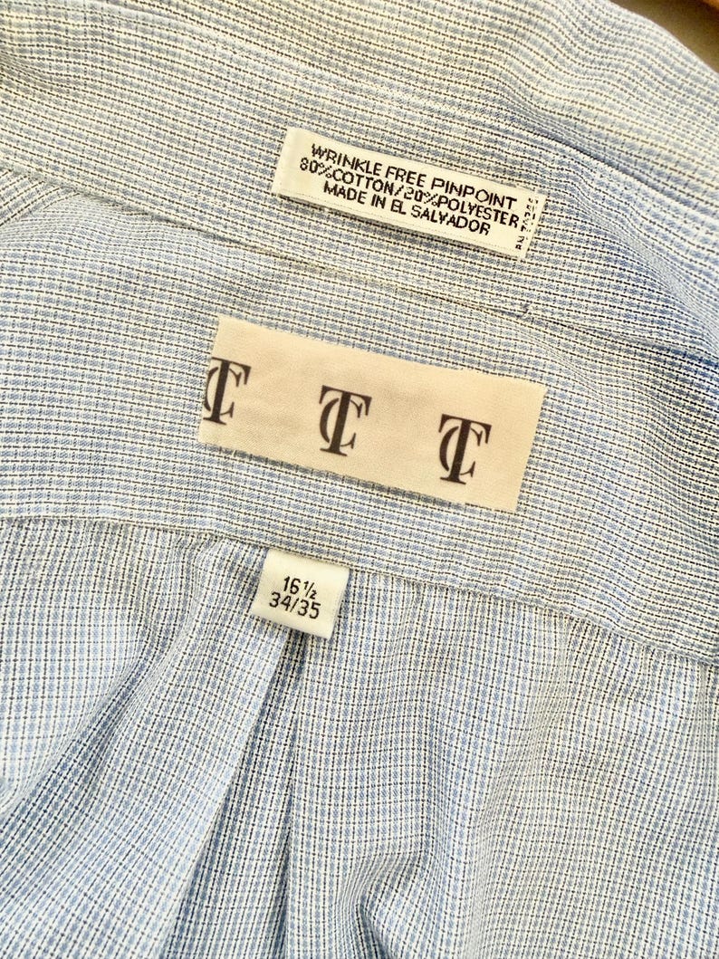 May include: Close-up of a light blue and white striped dress shirt. The shirt has a label that reads "WRINKLE FREE PINPOINT 80% COTTON/20% POLYESTER MADE IN EL SALVADOR." Another label displays the letters "TTT." A size label reads "16 1/2 34/35."