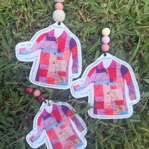 May include: Three hanging ornaments shaped like a patchwork coat with red, pink, and purple squares. Each ornament features the text "Momma sewed the love into every stitch of the coat. It didn't matter to me that we didn't have money. I felt rich because of the love that coat represented. It was a treasure beyond anything they could buy." and "Dolly Parton 1946-2026" with decorative beads.