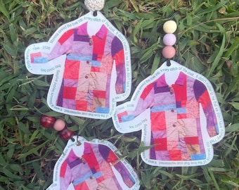 May include: Three hanging ornaments shaped like a patchwork coat with red, pink, and purple squares. Each ornament features the text "Momma sewed the love into every stitch of the coat. It didn't matter to me that we didn't have money. I felt rich because of the love that coat represented. It was a treasure beyond anything they could buy." and "Dolly Parton 1946-2026" with decorative beads.