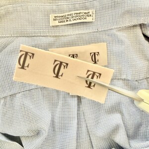 May include: Close-up of a light blue and white patterned shirt with a tag that reads "WRINKLE FREE PINPOINT 80% COTTON/20% POLYESTER MADE IN EL SALVADOR." Two beige tags with the letter "T" are being cut with scissors.