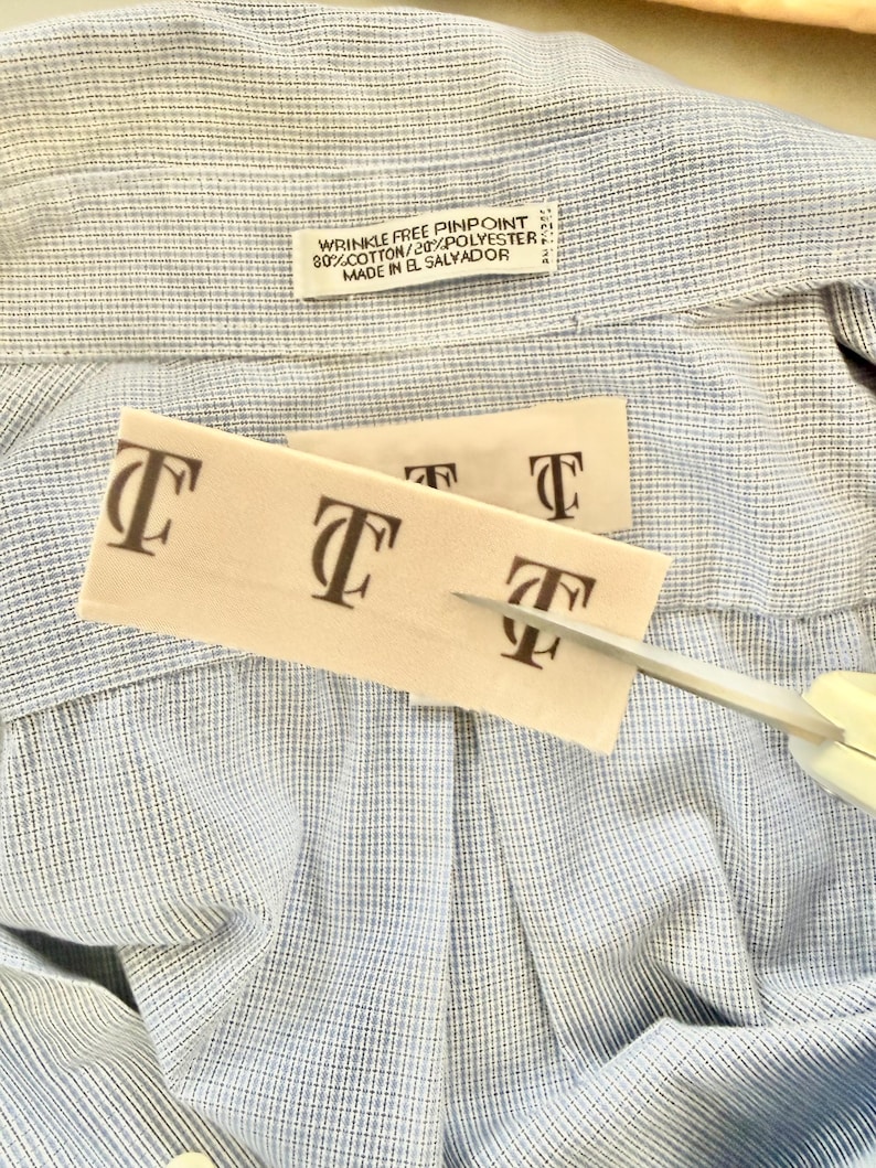 May include: Close-up of a light blue and white patterned shirt with a tag that reads "WRINKLE FREE PINPOINT 80% COTTON/20% POLYESTER MADE IN EL SALVADOR." Two beige tags with the letter "T" are being cut with scissors.
