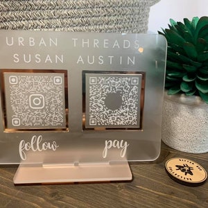 以下が含まれることがあります： 「URBAN THREADS SUSAN AUSTIN」というテキストと2つのQRコードが書かれた白いアクリル製の看板。左側のQRコードには「follow」、右側のQRコードには「pay」と書いてあります。