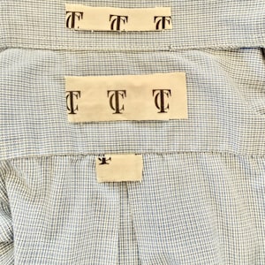 May include: Close-up of a light blue and white checkered shirt. The shirt has three small rectangular tags with the letter "T" printed on them. The tags are beige and are placed on the back of the shirt.