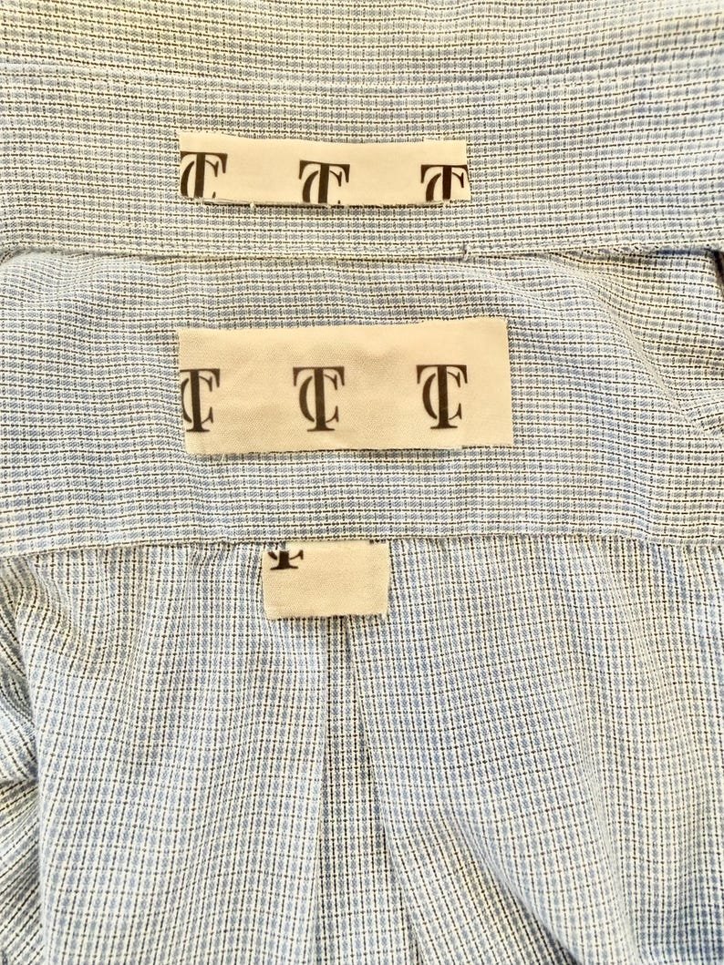 May include: Close-up of a light blue and white checkered shirt. The shirt has three small rectangular tags with the letter "T" printed on them. The tags are beige and are placed on the back of the shirt.